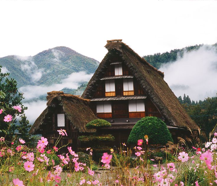 a house with thatched roof and pink flowers in the foreground, surrounded by mountains