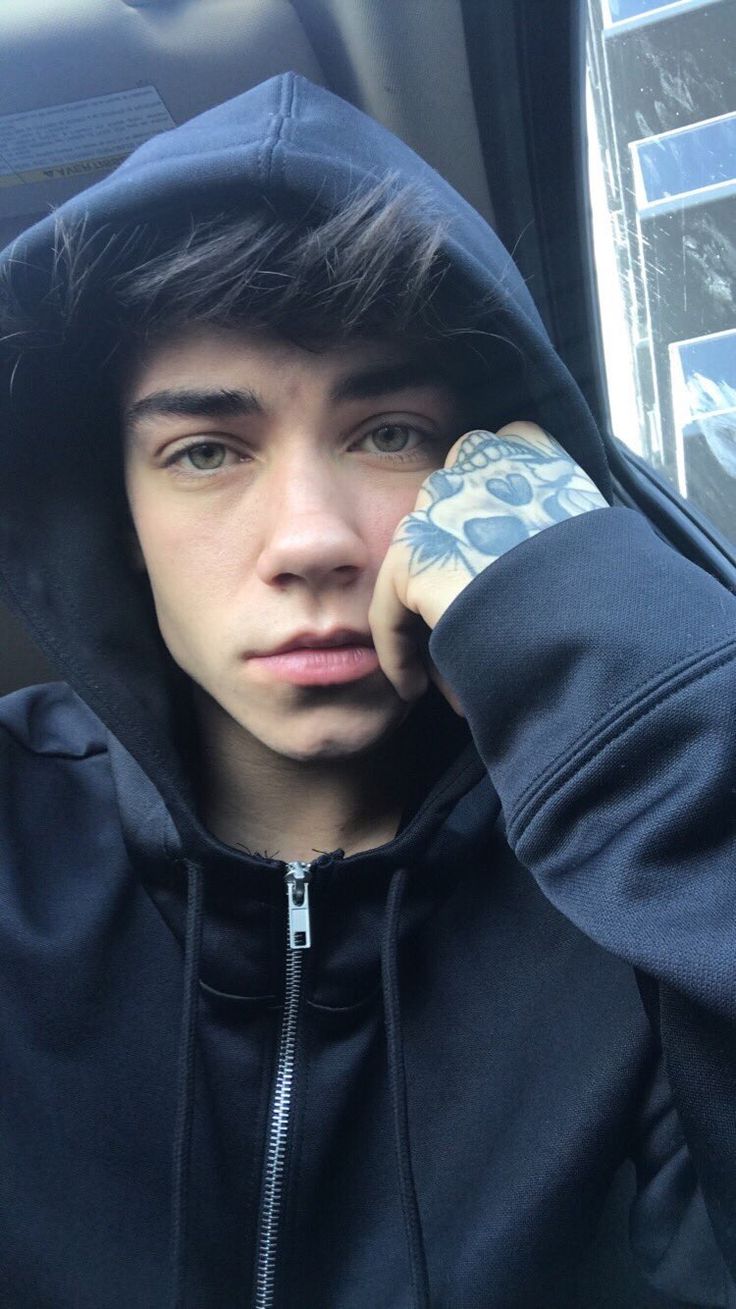 a young man in a black hoodie with tattoos on his arm and hand near his face