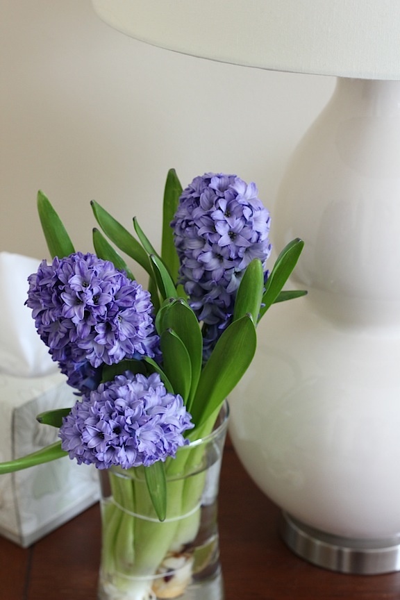 purple flowers are in a glass vase on a table next to a white lamp shade