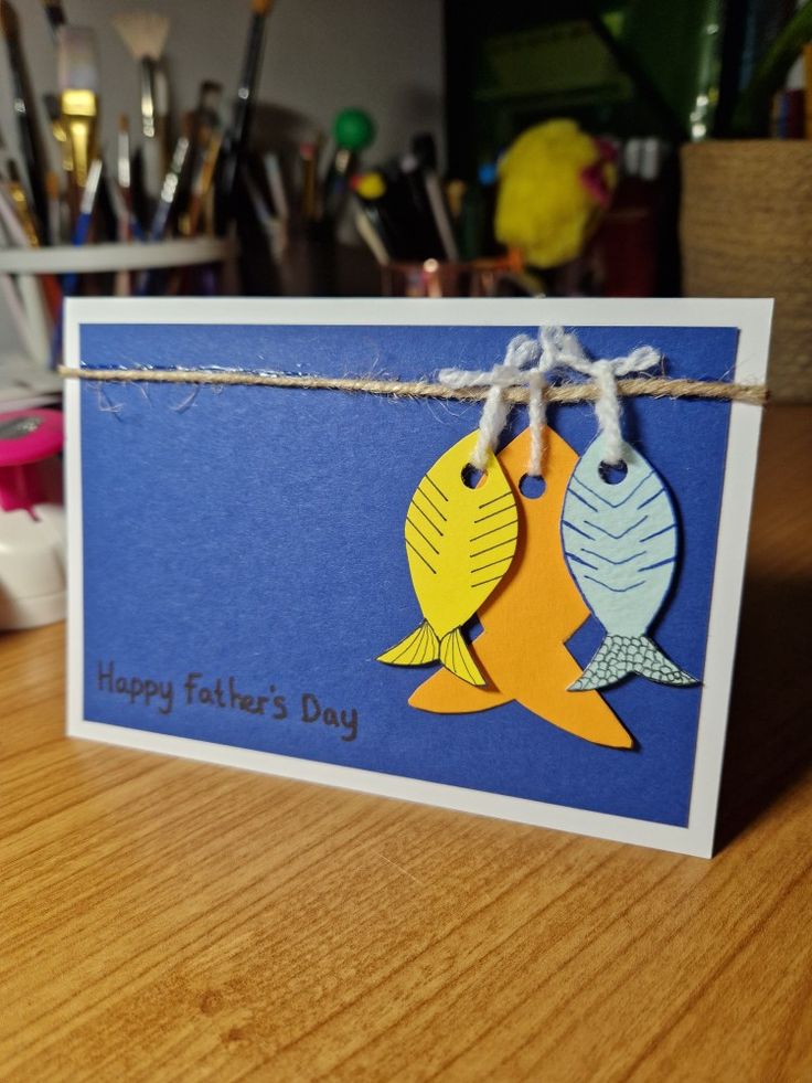 a card with two fish hanging from it's side on a wooden table next to paintbrushes