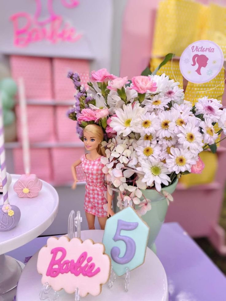 there is a birthday cake with flowers in the vase and a doll next to it