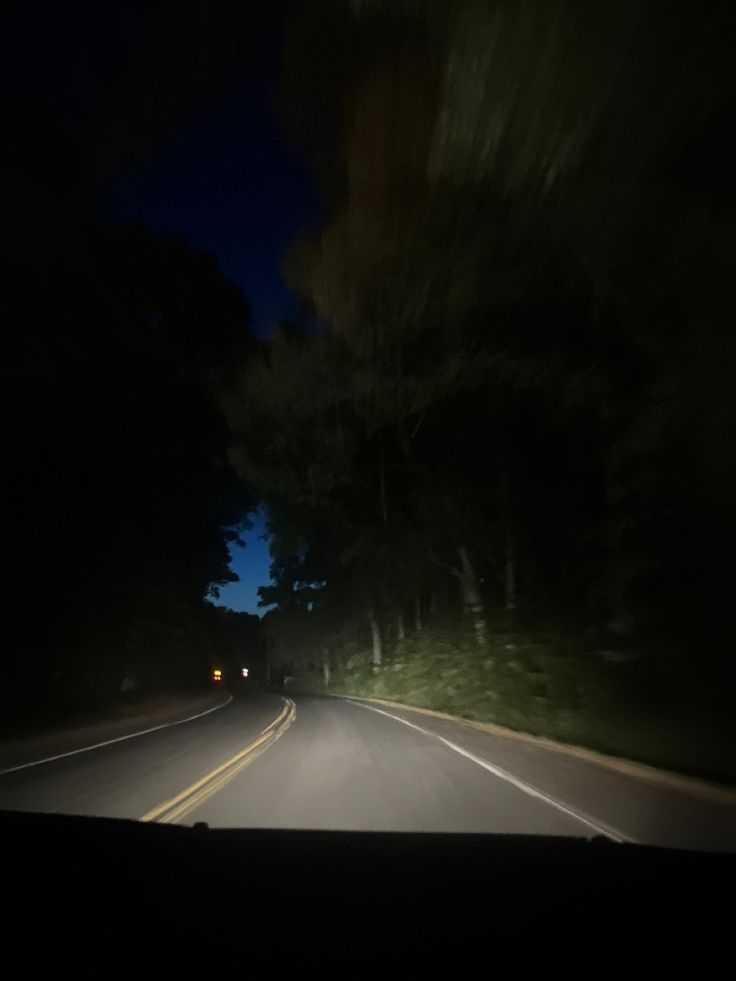 a car driving down the road at night with its headlights on and trees in the background