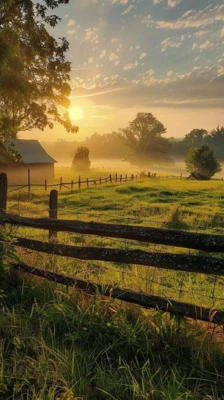 the sun is setting over a farm field with a fence and barn in the distance