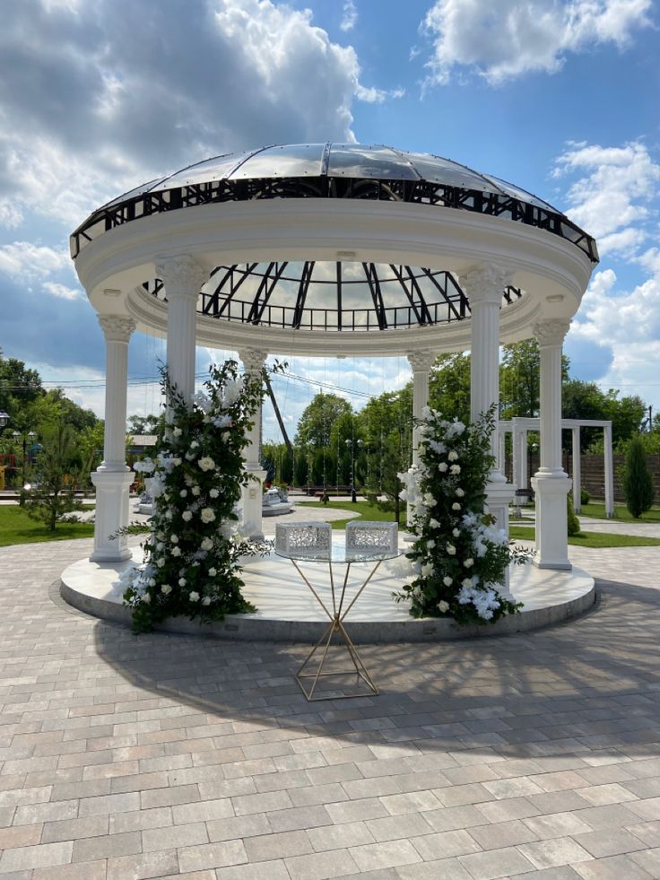 a gazebo with white flowers and greenery on the sides under a cloudy blue sky