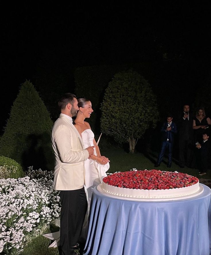 a bride and groom standing in front of a large cake with berries on it at night