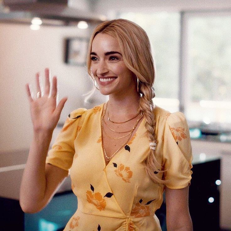 a woman in a yellow dress waving at the camera with her hand up and smiling