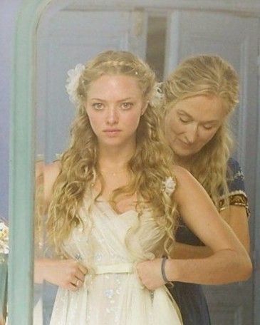 two women are looking in the mirror and one is wearing a white dress with flowers on her head