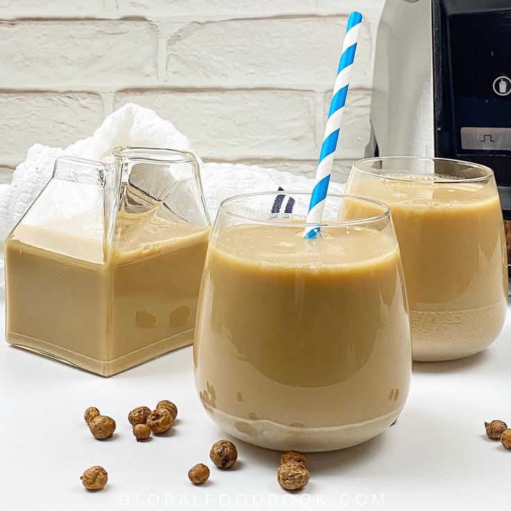 two glasses filled with peanut butter milk next to a blender and some nuts on the table