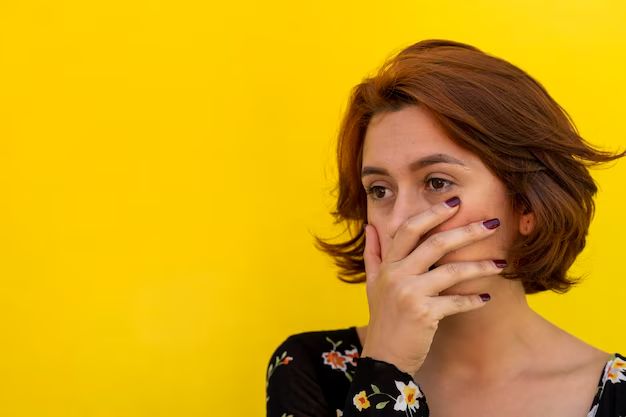 a woman covers her face with her hands while standing against a yellow wall, looking at the camera