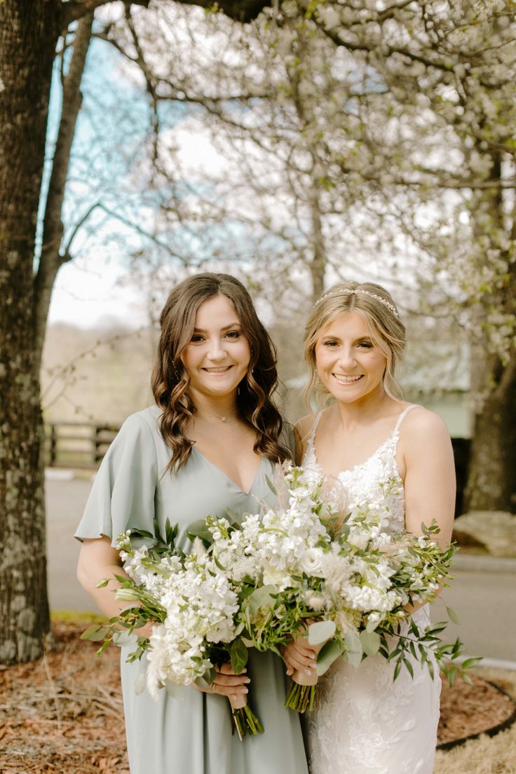 two bridesmaids pose for a photo in front of trees and flowers at their wedding