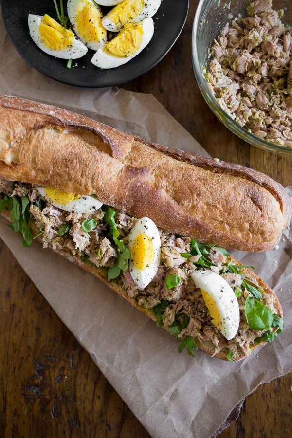 a sandwich with meat, eggs and greens sits on wax paper next to a bowl of rice