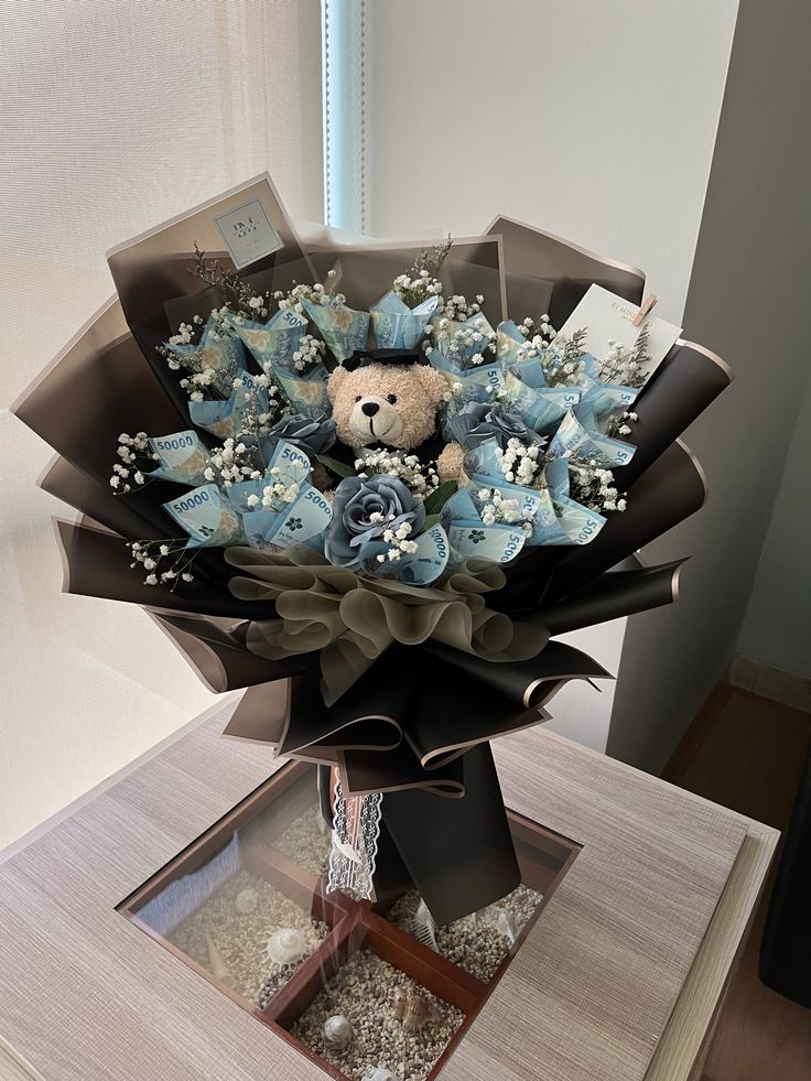 a bouquet of flowers with a teddy bear in the center on a table next to a window