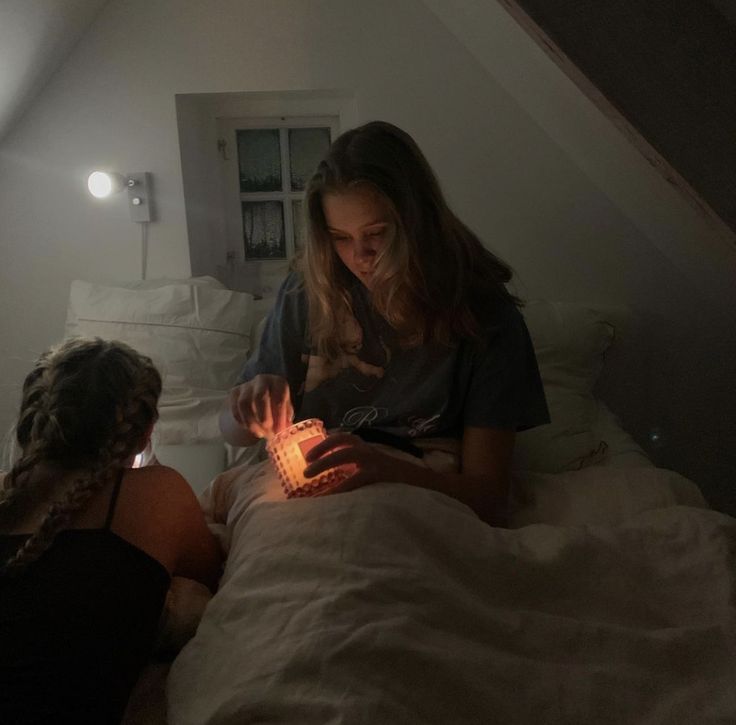 two women sitting on a bed looking at something in the dark with one woman holding a lit candle