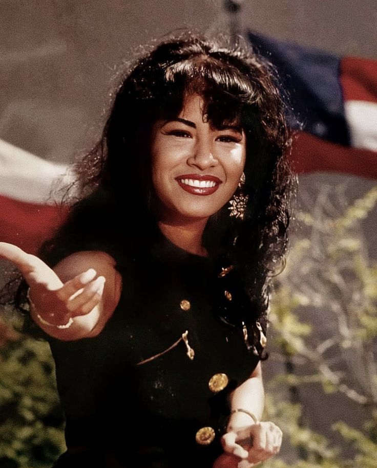 a woman with long black hair pointing to the side and smiling at the camera, in front of an american flag