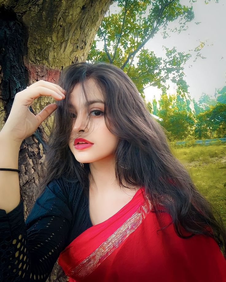 a woman in a red and black sari standing next to a tree with her hand on her head