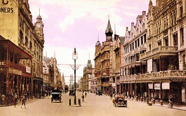 an old time photo of people walking and riding in the street with buildings on either side