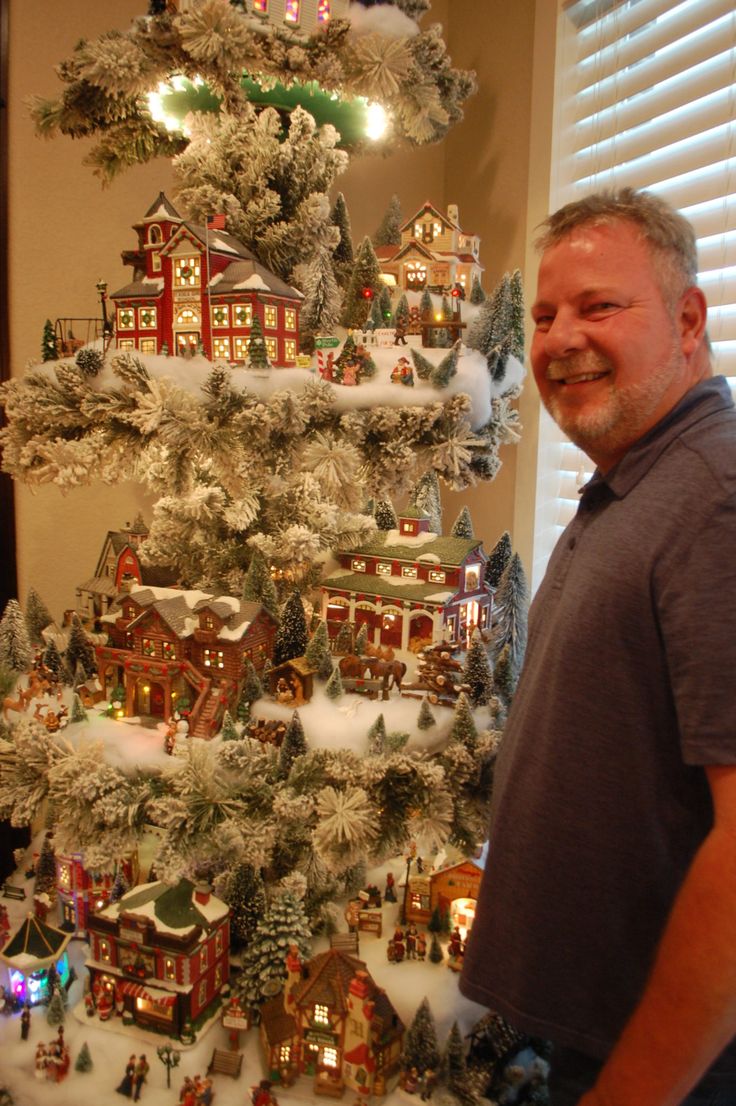 a man standing in front of a christmas tree that has snow on it and houses