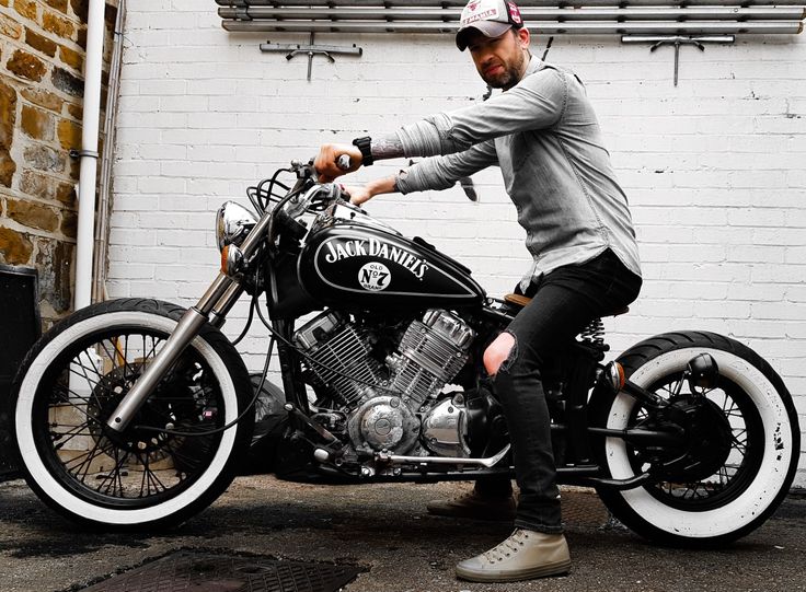 a man sitting on top of a black motorcycle next to a white brick building and holding onto the handle bars