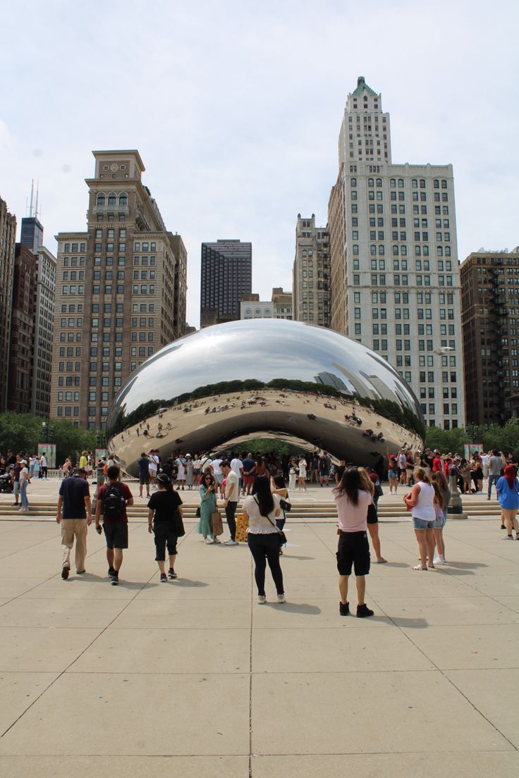 many people are walking around in front of the chicago beanhead sculpture and skyscrapers