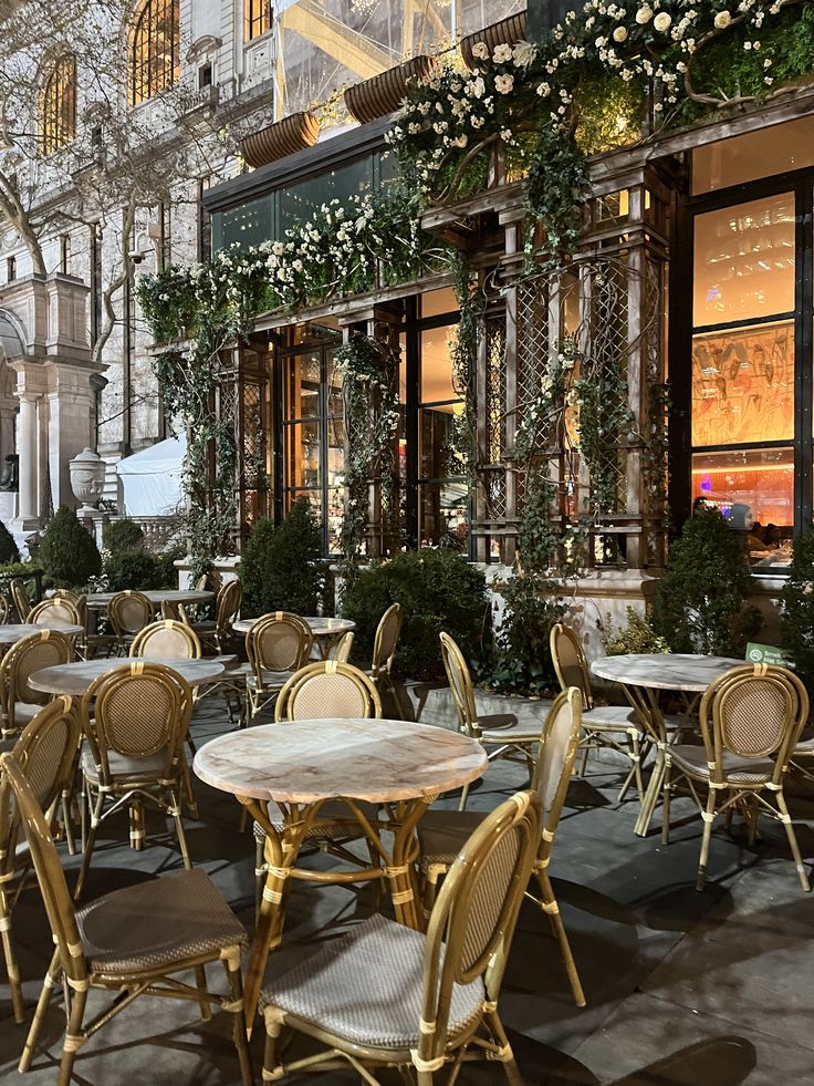 tables and chairs are set up in front of a building with flowers on the windows