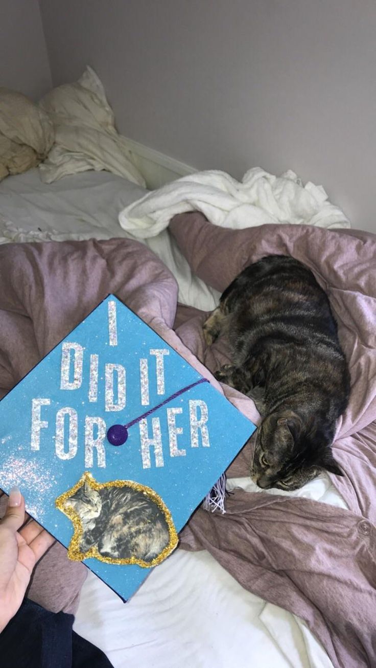 a cat laying on top of a bed next to a blue graduation cap that says i did it for her