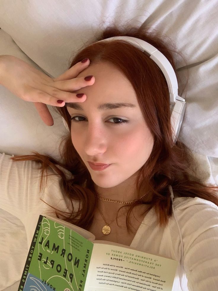 a woman laying in bed while reading a book and listening to headphones on her ears