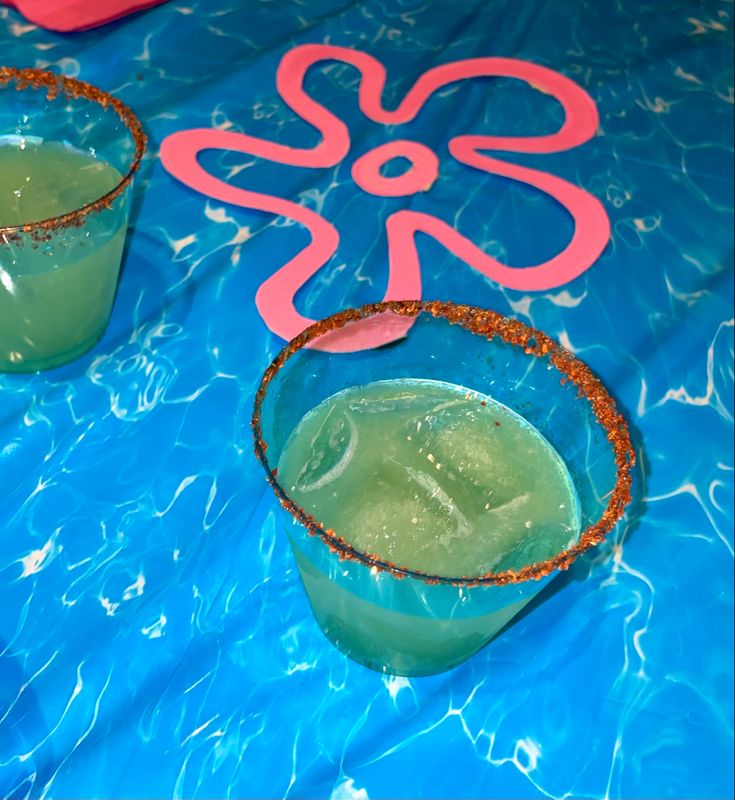 two glasses filled with green liquid sitting on top of a blue table covered in water