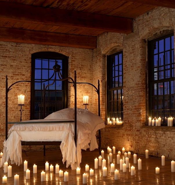 a bed with lots of lit candles in front of two windows and a brick wall