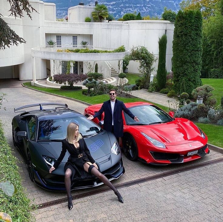 a man and woman sitting on top of two sports cars in front of a mansion