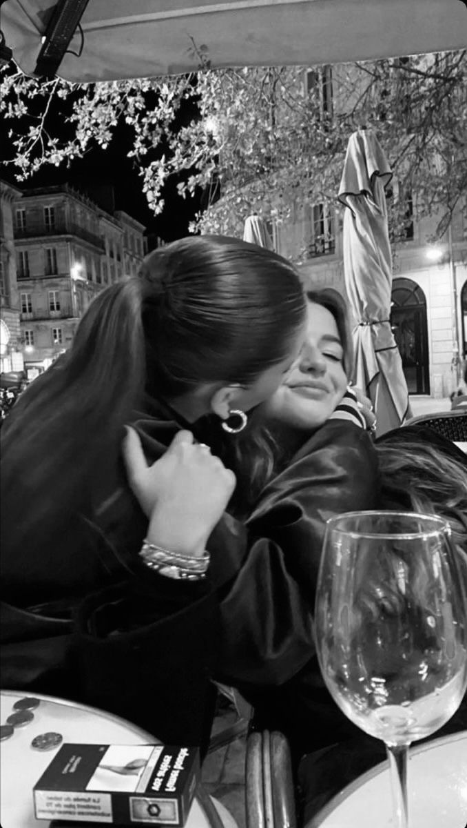 two women hugging each other at an outdoor table with wine glasses and umbrellas in the background