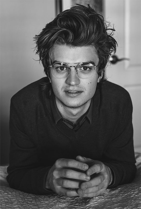 a black and white photo of a man with glasses laying on a bed looking at the camera