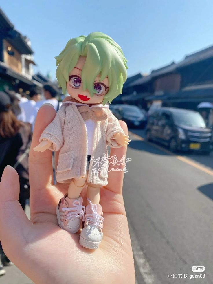 a hand holding a small doll in the middle of a street with cars parked on both sides