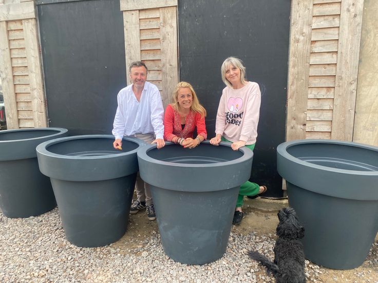 two people and a dog are sitting in front of large plastic planters on the ground