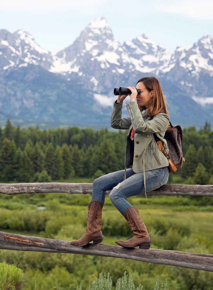 a woman sitting on top of a wooden fence looking into the distance with mountains in the background