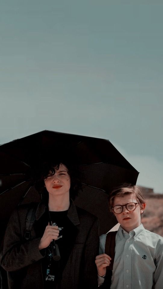 two young men standing under an umbrella in the rain, one with glasses on his head