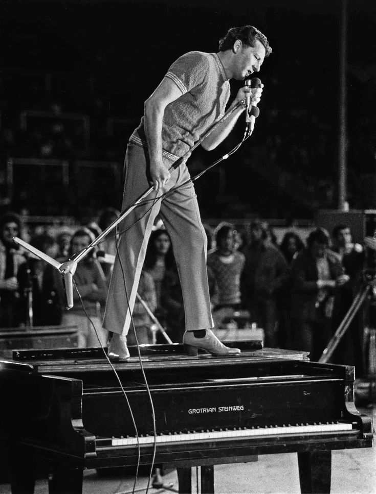 a man standing on top of a piano in front of a crowd with microphones