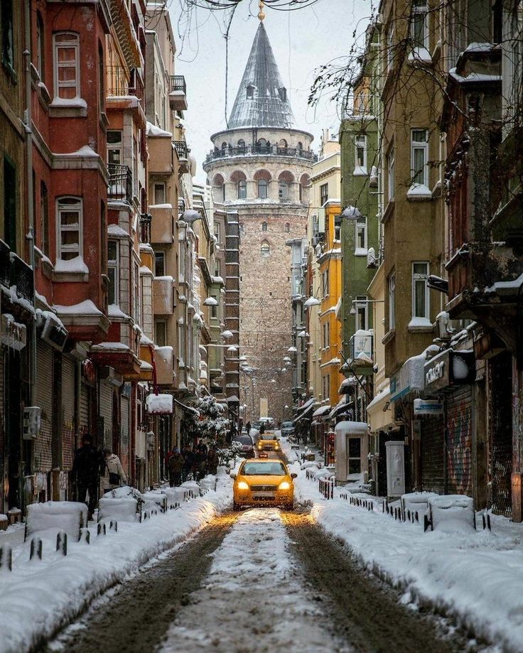 a yellow taxi cab driving down a snow covered street in front of tall buildings with a clock tower