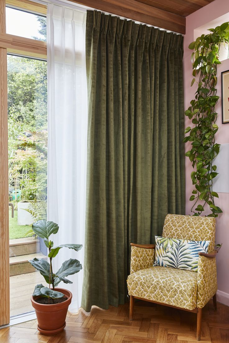 a living room with green curtains and a chair in front of a window filled with potted plants