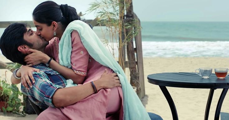 a man and woman kissing on the beach next to an ocean side table with drinks in front of them