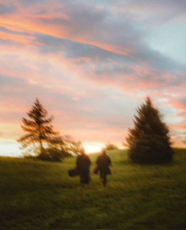 two people are walking in the grass at sunset with trees and clouds behind them as the sun sets