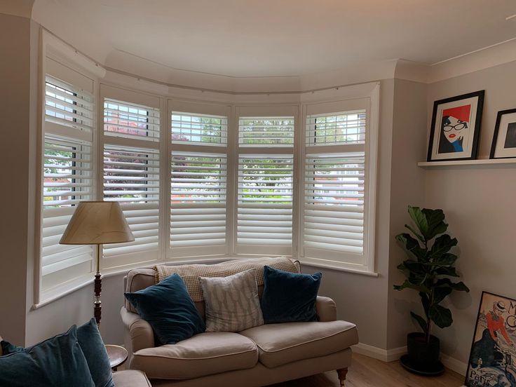 a living room filled with furniture and windows covered in white shutters, next to a lamp