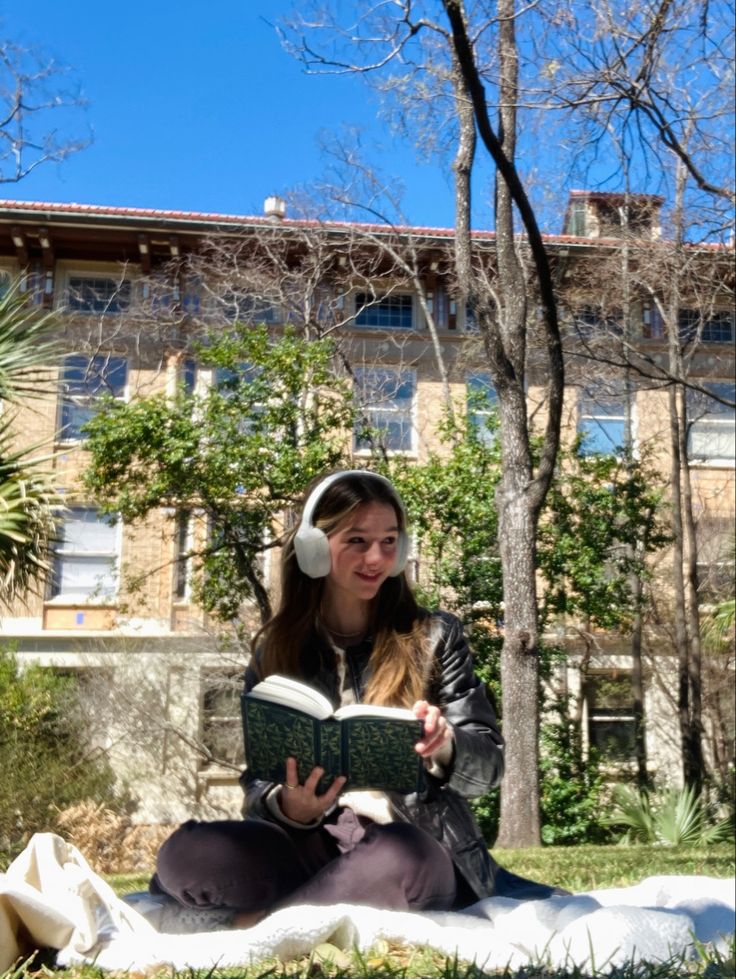 a woman sitting on the ground reading a book and listening to headphones in front of a building