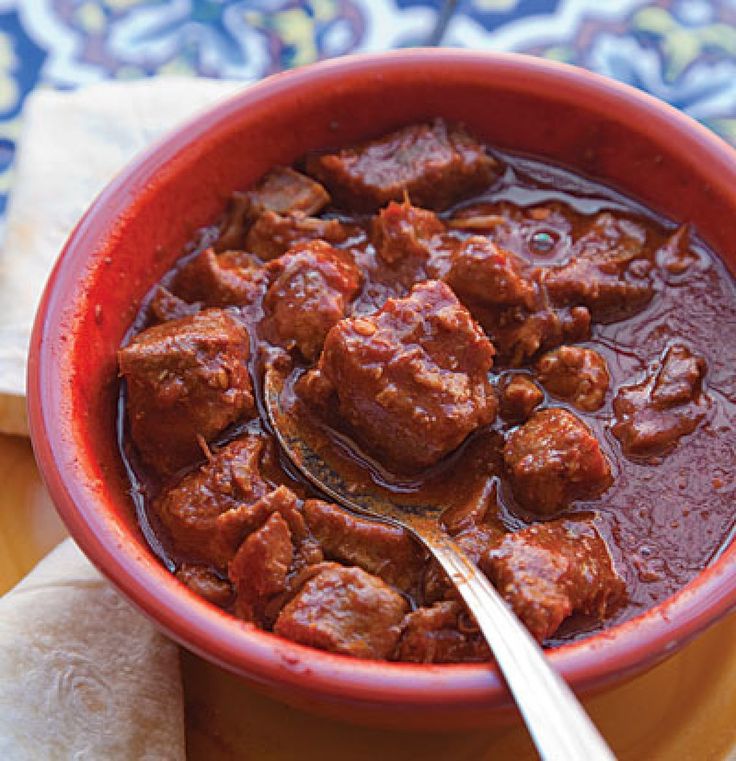 a red bowl filled with meat and sauce on top of a wooden table next to a napkin