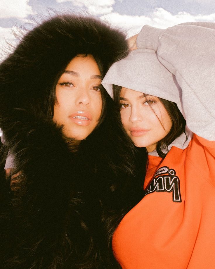 two young women posing for a photo in front of a snow covered background, one wearing an orange hoodie and the other with black fur on her head