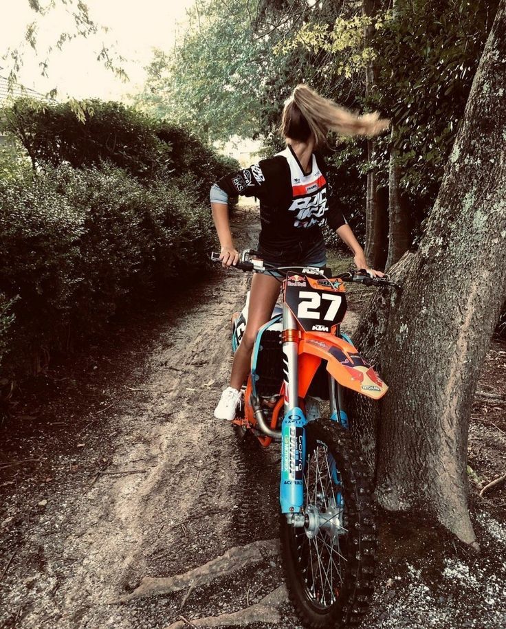 a woman riding on the back of a dirt bike down a forest trail next to a tree