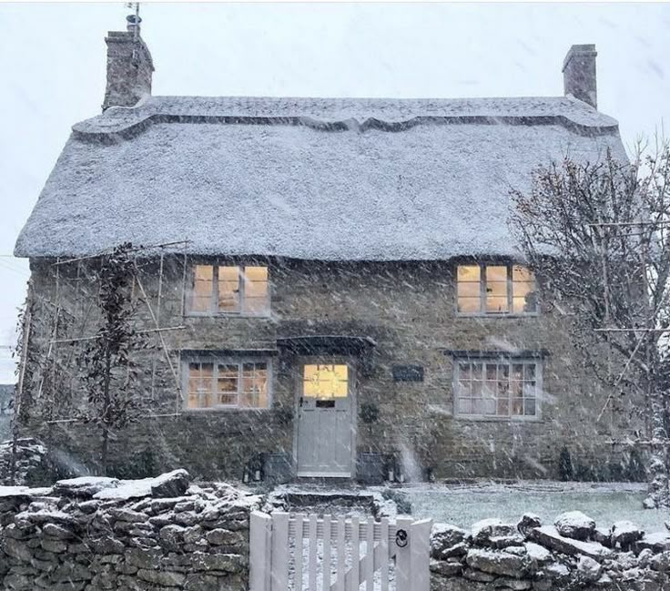 a stone house covered in snow next to a fence and gate with lights shining through the windows