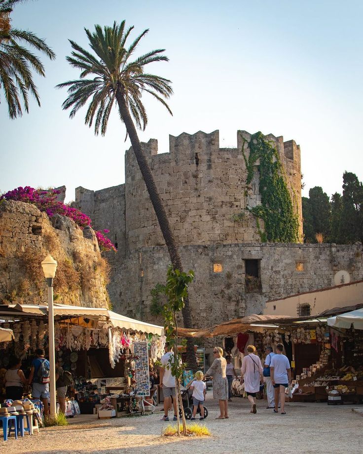 people are walking around an outdoor market in front of a stone castle with palm trees