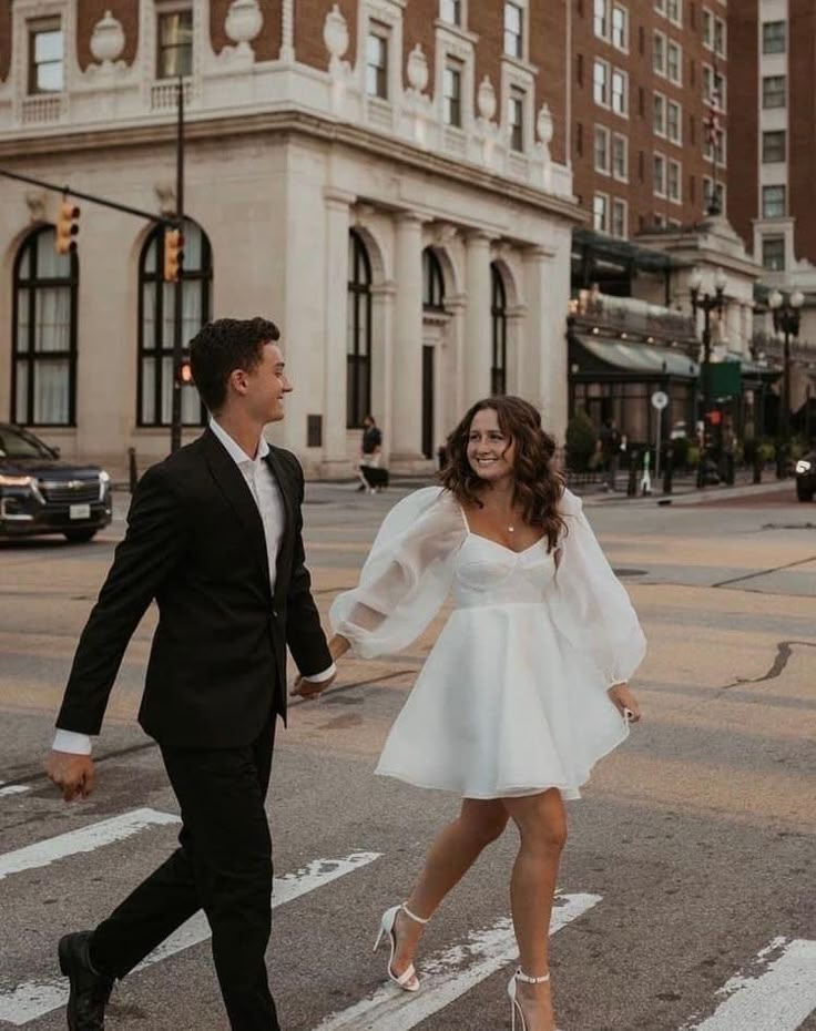 a newly married couple crossing the street in front of a large building at sunset, dressed in black and white