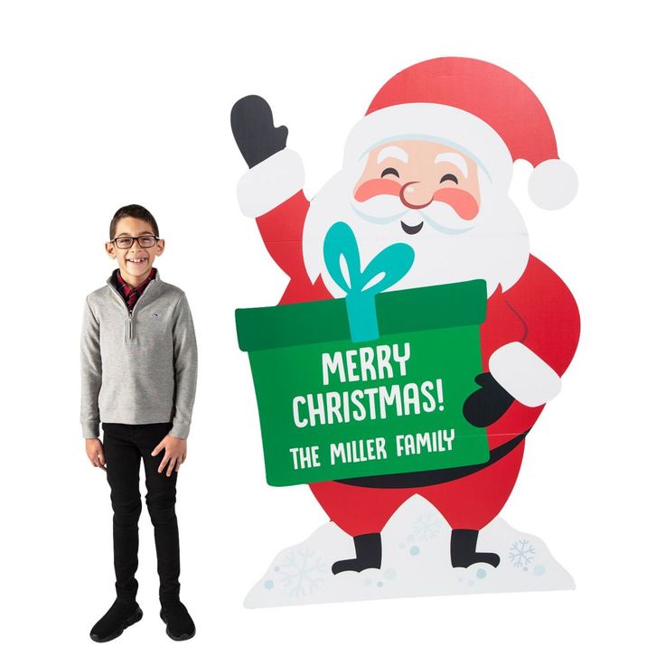 a young boy standing next to a large christmas sign with santa holding a green box