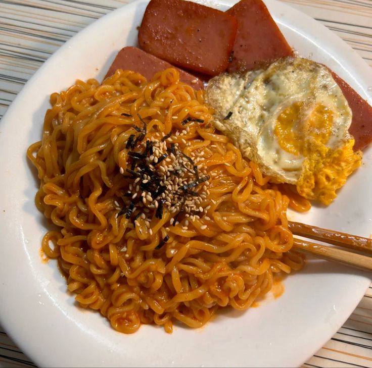 a white plate topped with noodles, meat and an egg next to chopsticks